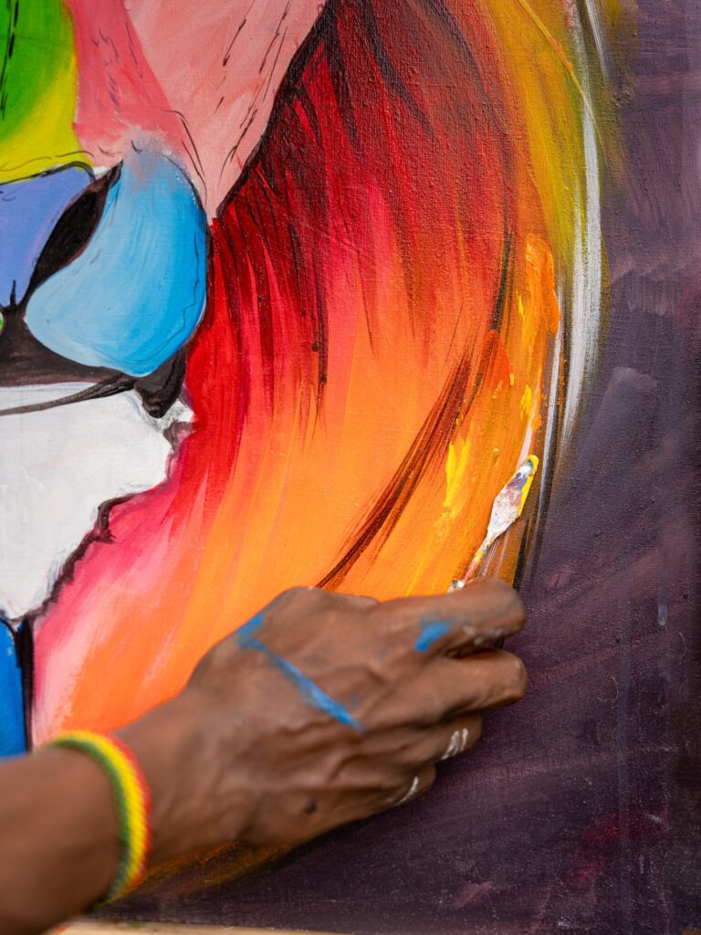 Close-up of a person's hand painting a vibrant abstract scene on a canvas. The artwork features bright hues of orange, red, yellow, green, and blue. The painter's hand, with traces of paint, holds a brush. A colorful striped wristband adorns their wrist.