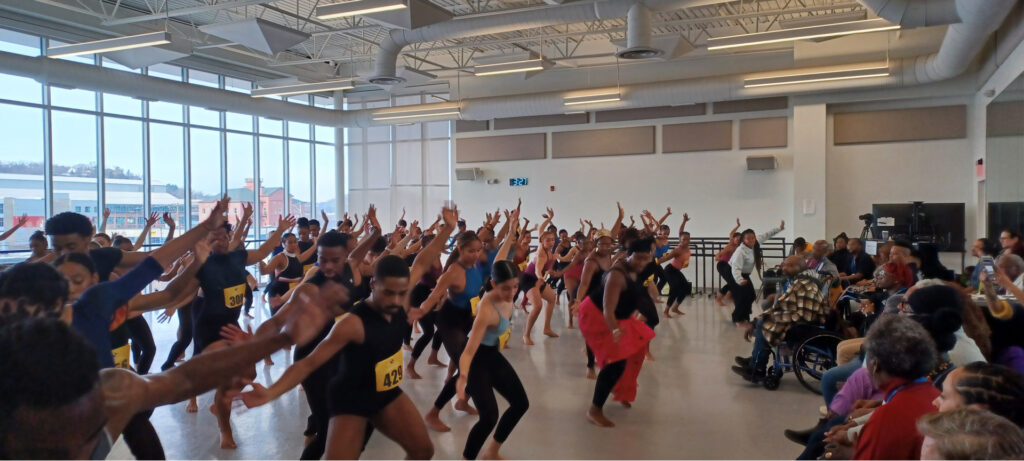 A large group of dancers in a rehearsal studio practices with energetic arm movements. Each wears a numbered tag. People seated on the right observe. The spacious room has large windows and a high ceiling. The atmosphere is lively and focused.