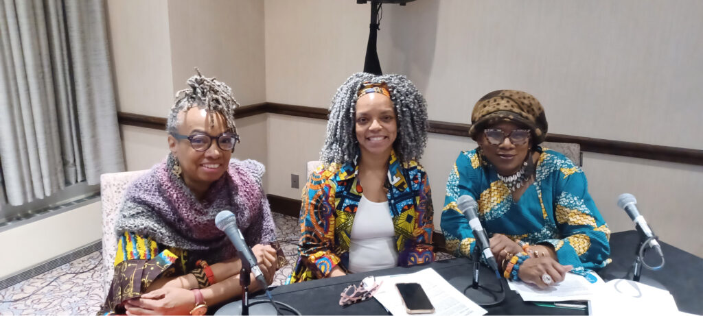 Three people sit at a table with microphones in front of them, smiling. They wear colorful, patterned clothing with distinct headwear and accessories. Papers and a phone are on the table. The background features a beige wall and a curtain on the left side.