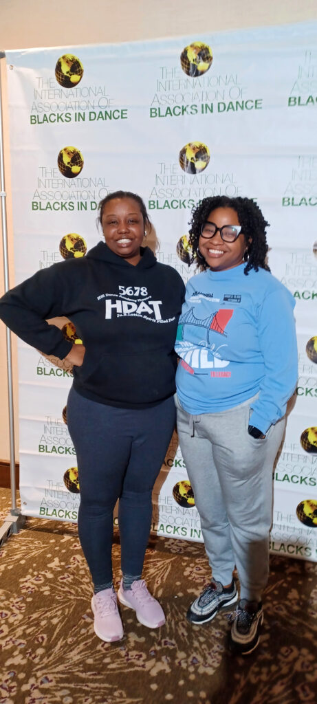 Two people stand smiling in front of a banner that reads "The International Association of Blacks in Dance." One is wearing a black sweatshirt and gray pants, the other a blue sweatshirt and gray joggers. The background is a patterned carpet.