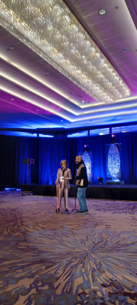 Two people stand on a carpeted stage, speaking into microphones. The person on the left wears a striped suit and hat, holding a wooden cane. The person on the right wears a dark jacket with gold stripes. A backdrop with geometric patterns is behind them, under a ceiling with bright, decorative lights.