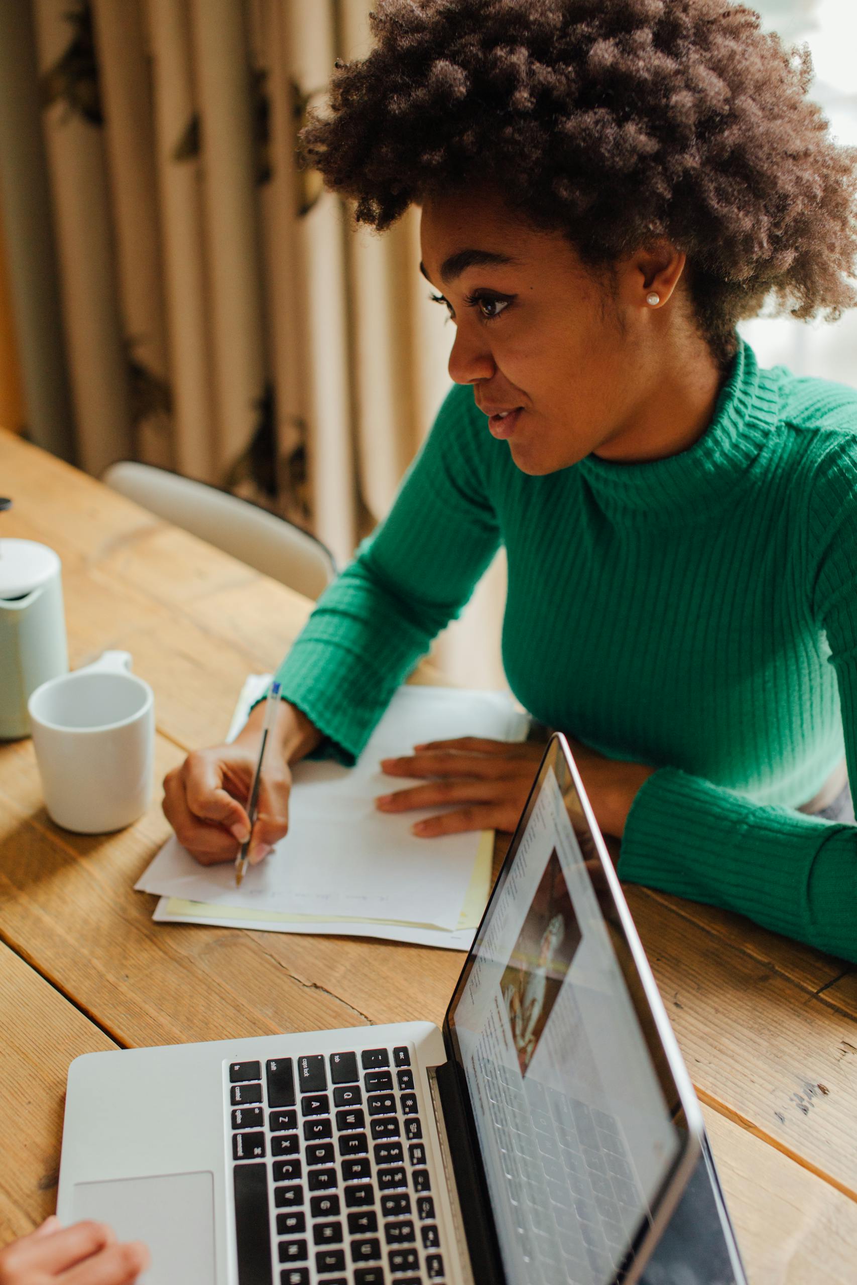 A young woman with afro hair writing in a notebook while using a laptop indoors.