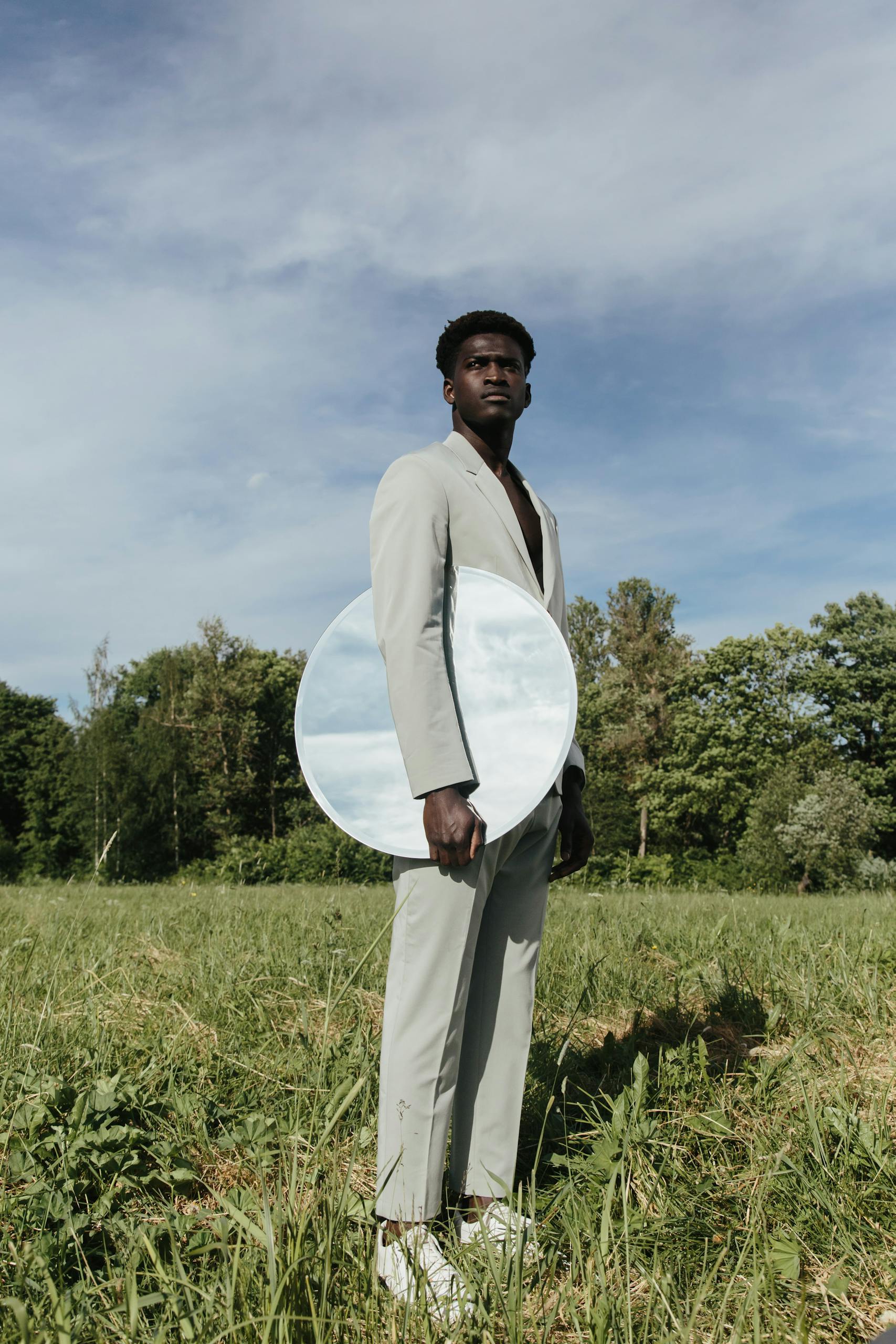 African American man in a suit holding a round mirror outdoors, reflecting sky and landscape.