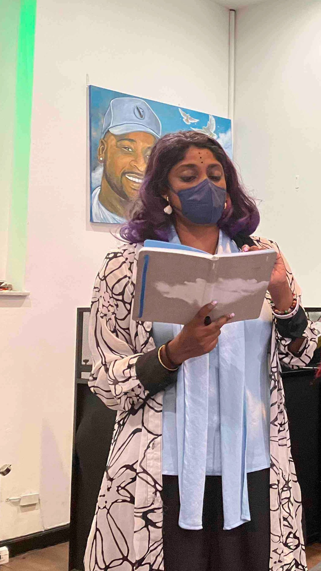 A woman with wavy purple hair, wearing a patterned coat, light blue scarf, and face mask, reads from a notebook. Behind her, a portrait of a smiling man in a baseball cap hangs on a white wall. The scene is indoors with soft lighting.