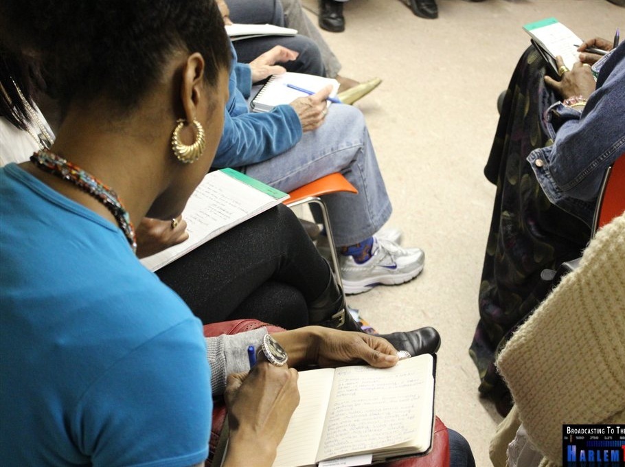 A woman with braided hair and gold hoop earrings writes in a notebook on her lap. She sits among a group of people, all holding notebooks or writing pads. The setting appears to be a classroom or community meeting. The floor is beige and chairs are orange.