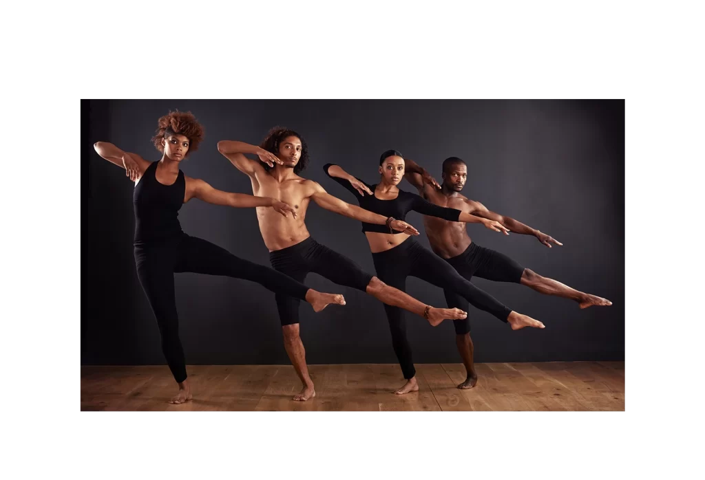 Four dancers in black attire pose in a line against a dark background, raising their left legs and extending their right arms forward and left arms back, creating a dynamic, synchronized formation on a wooden floor.